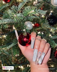 Long Silvery White Nails With Red Glitter And Rhinestones Holding Cjhristmas Decoration In Front Of A Christmas Tree