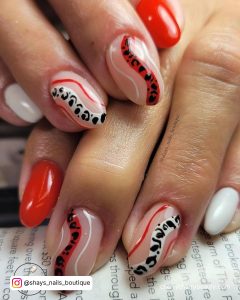 Short Oval Red Black And White Nails With Swirly White, Black, Red, And Black Cow Print Design On The Middle And Ring Fingers Placed On A Book