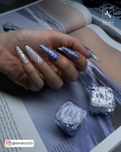 Hand With Almond Shaped White Chrome Nails With Glitter On A Opened Book