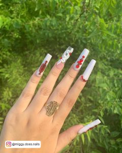 Long Red And White Acrylic Nails With Bloody Red Rhinestones And Flower Design Over Green Grass Background