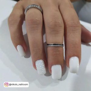 Milky Nails With White Glitter On Ring Finger Placed Over A White Shelf