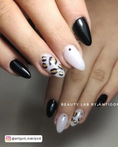 Oval Black And White Cow Print Nails With Plain Black And White Polish On The Index, Pinky, And Middle Finger, And Gold And Black Cow Print On The Ring Finger.