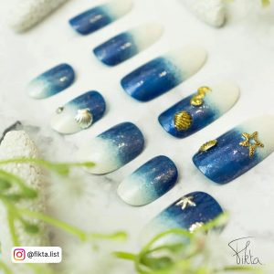Shimmery Ombre Nails Blue And White With Gold Star And Clam Designs On White Surface