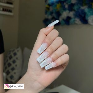 White French Tip Nails With Glitter Infront Of A Floral Wall Hanging