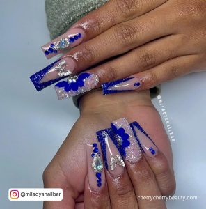Bling Acrylic Royal Blue Nails With Flowers, Glitter, Rhinestones, And Butterfly Over White Surface