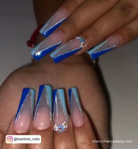Royal And Light Blue Tip Acrylic Nails With Glitter And Flowers Over White Surface